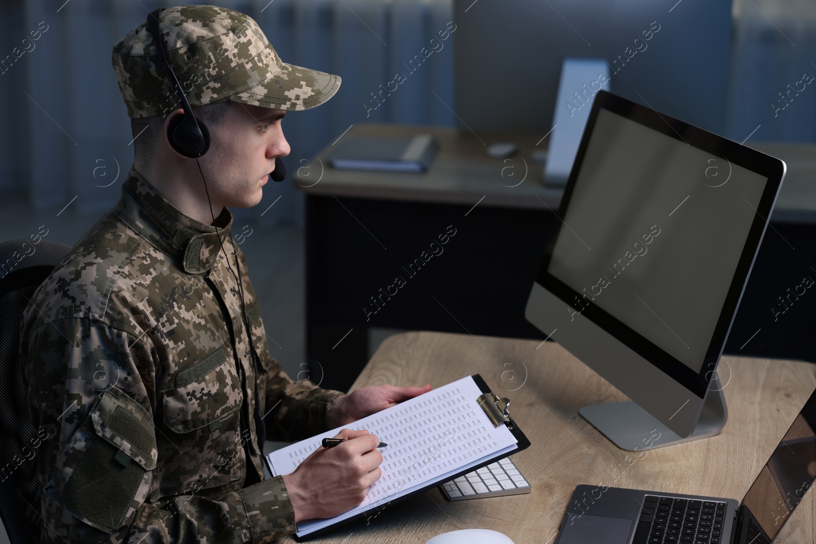 Photo of Military service. Soldier with clipboard and headphones working at table in office