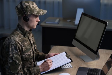 Photo of Military service. Soldier with clipboard and headphones working at table in office