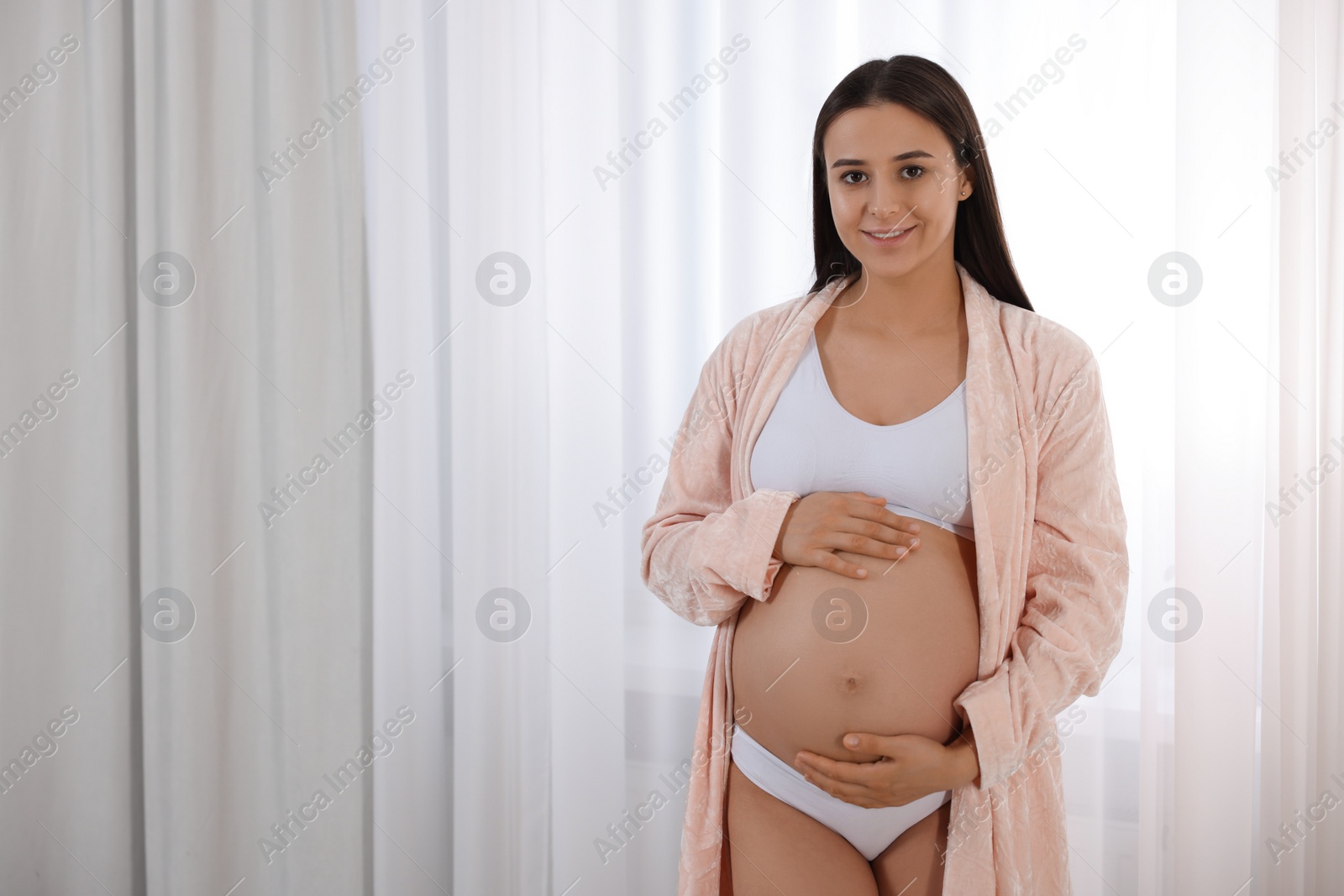 Photo of Beautiful pregnant woman in stylish comfortable underwear and robe indoors, space for text