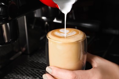 Photo of Barista pouring steamed milk from pitcher into glass cupfresh aromatic coffee in cafe, closeup