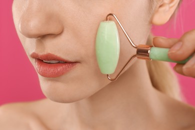 Photo of Young woman using natural jade face roller on pink background, closeup