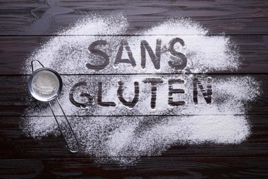 Photo of Sifter and phrase Gluten free written in French with flour on dark wooden table, flat lay