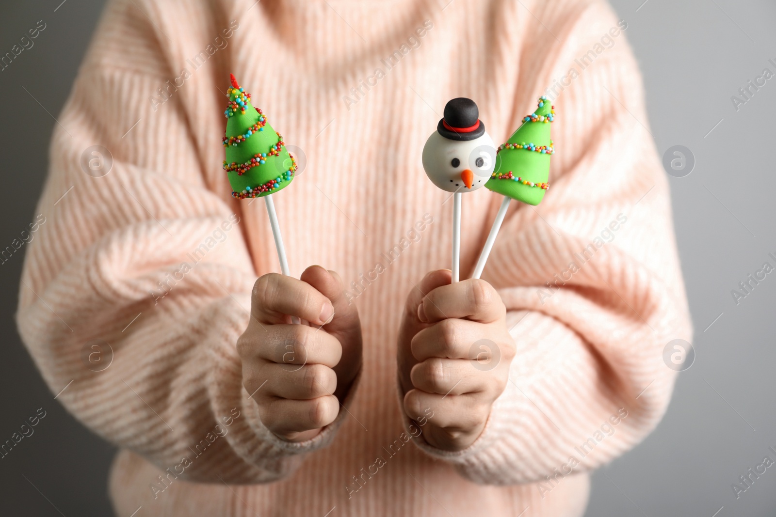 Photo of Woman holding delicious Christmas themed cake pops on grey background, closeup