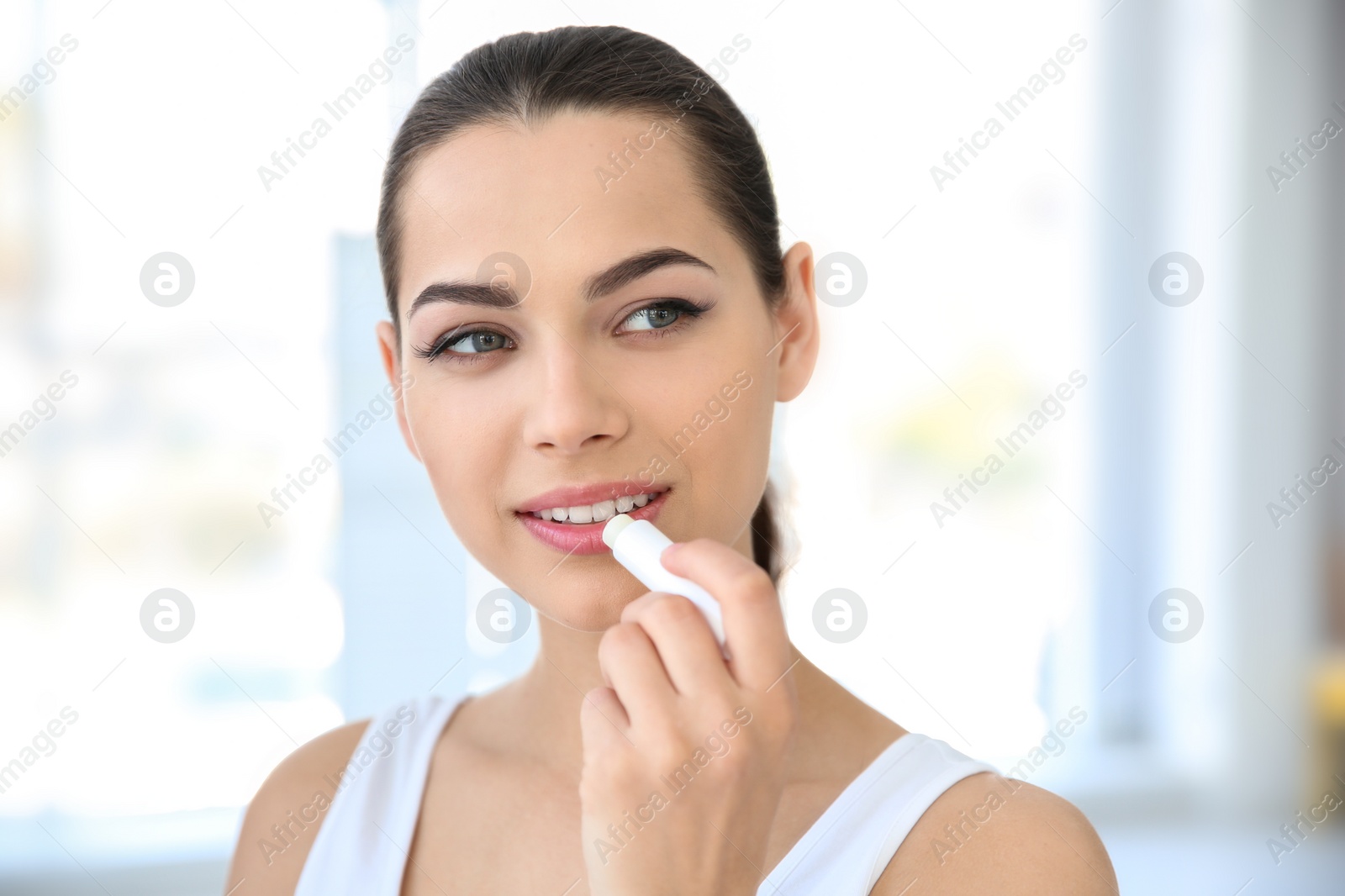 Photo of Young woman applying balm on her lips indoors