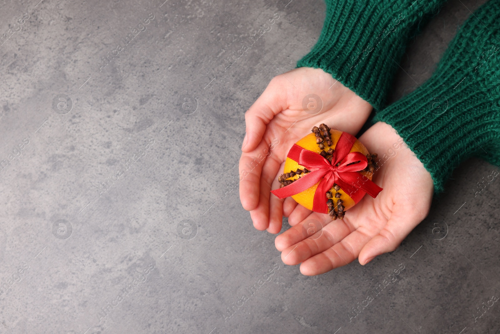 Photo of Woman holding pomander ball with red ribbon made of fresh tangerine and cloves at grey table, top view. Space for text