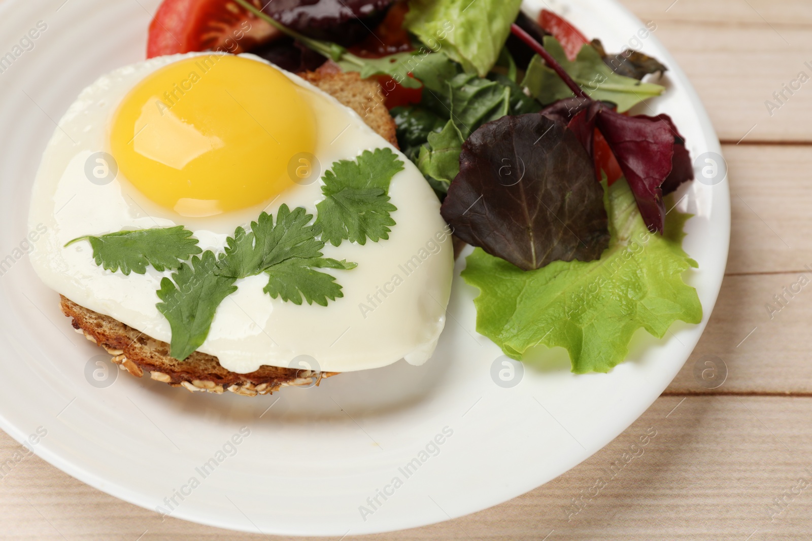 Photo of Plate with tasty fried egg, slice of bread and salad on light wooden table, closeup