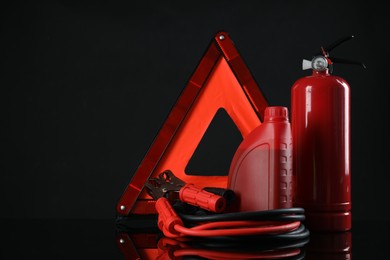 Photo of Emergency warning triangle, red fire extinguisher, battery jumper cables and motor oil on black background, space for text. Car safety