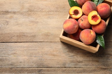 Photo of Cut and whole fresh ripe peaches with green leaves in crate on wooden table, above view. Space for text