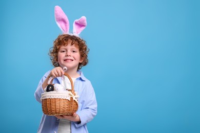 Photo of Portrait of happy boy in cute bunny ears headband holding wicker basket with Easter eggs on light blue background. Space for text