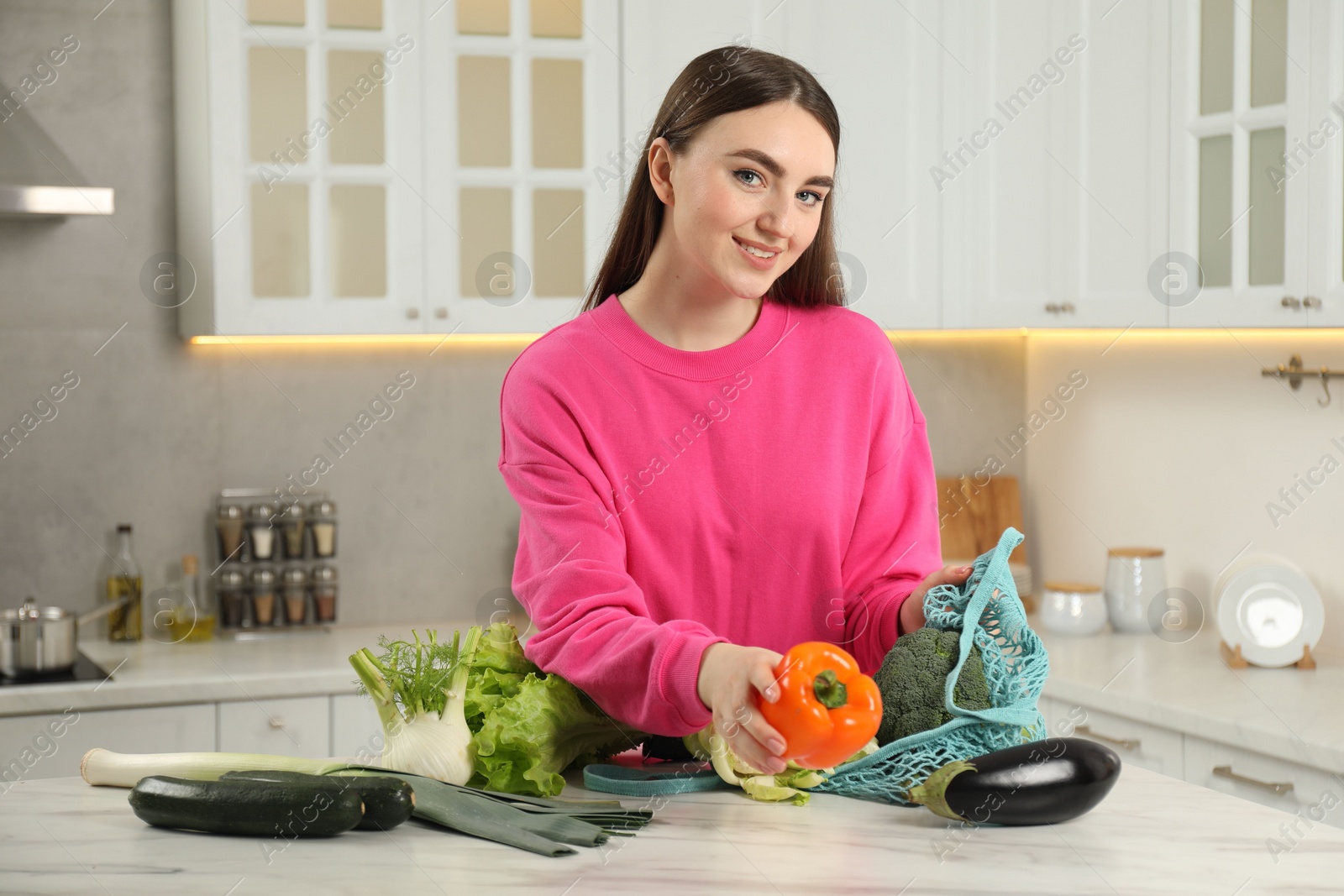 Photo of Woman taking pepper out from string bag at light marble table in kitchen