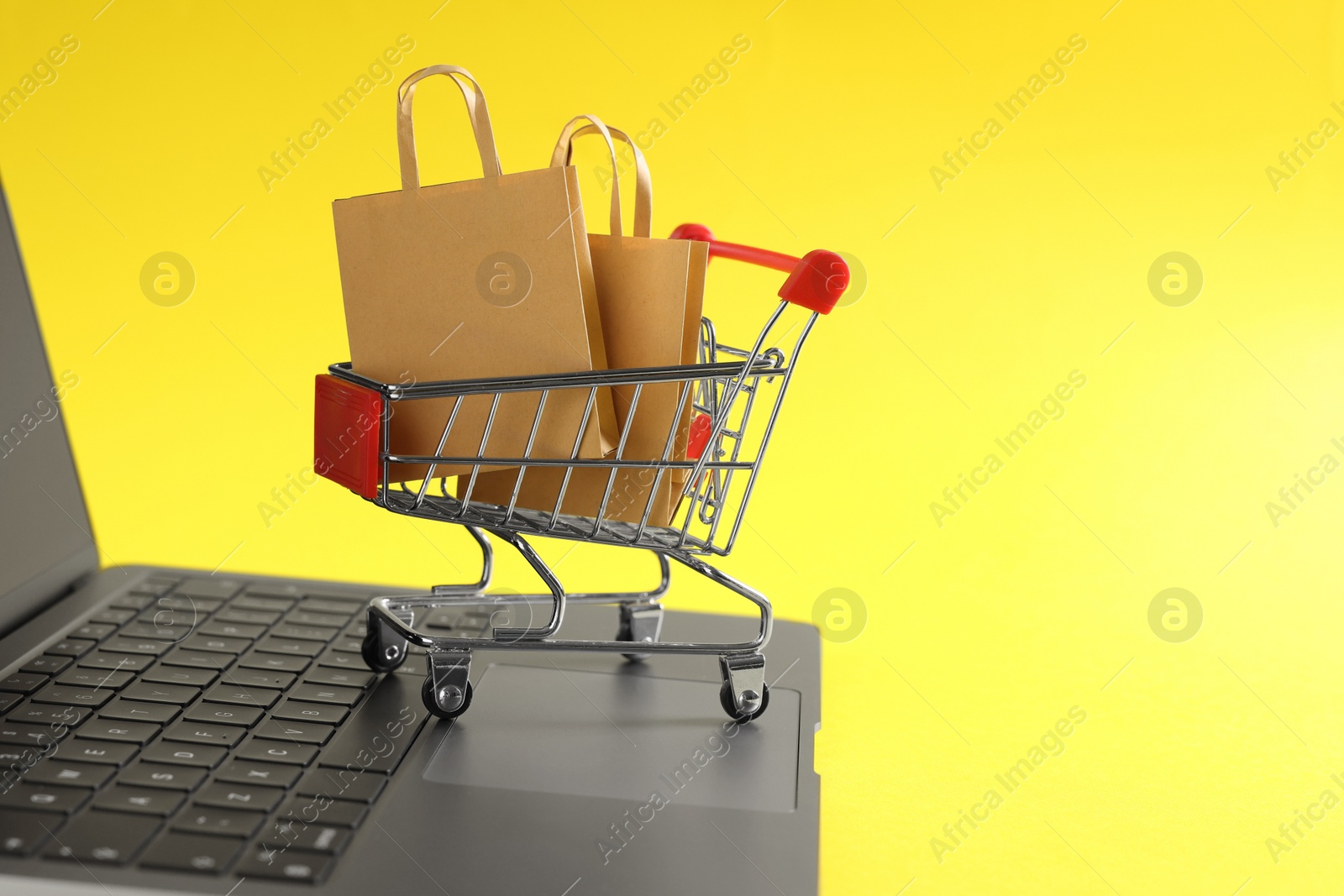 Photo of Online store. Laptop, mini shopping cart and purchases on yellow background, closeup. Space for text