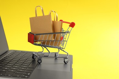 Photo of Online store. Laptop, mini shopping cart and purchases on yellow background, closeup. Space for text