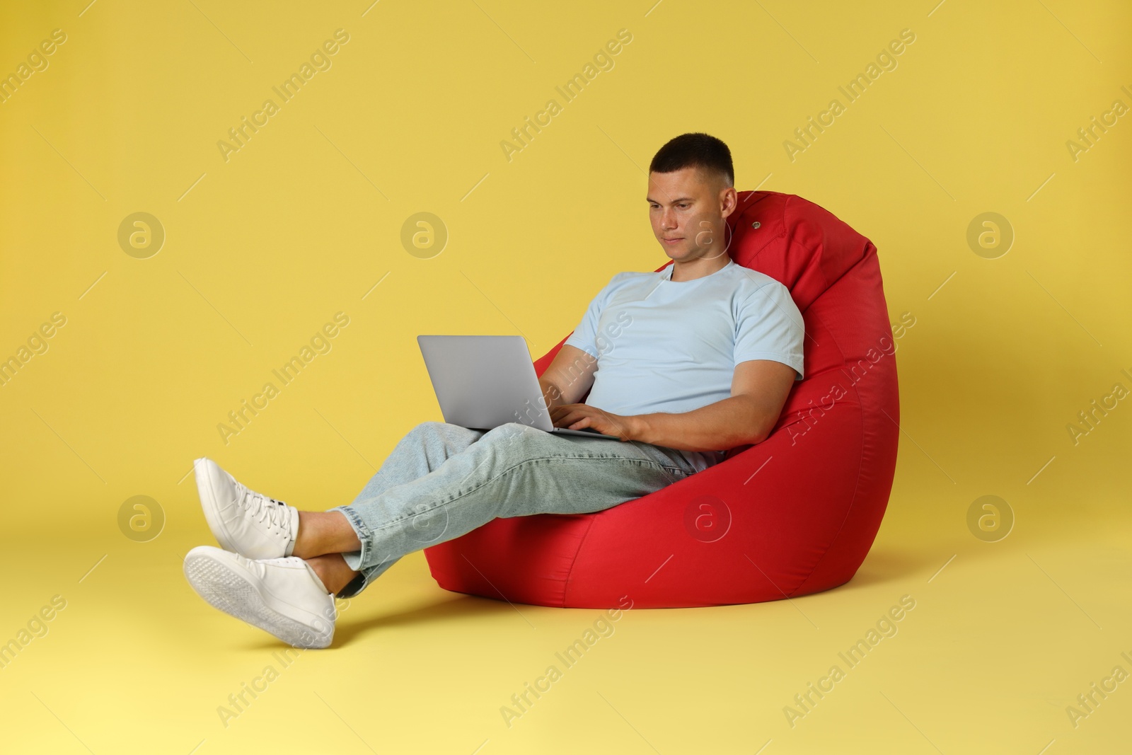 Photo of Handsome man with laptop on red bean bag chair against yellow background