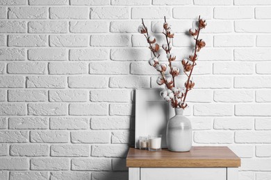 Photo of Cotton branches with fluffy flowers in vase on wooden table near white brick wall. Space for text