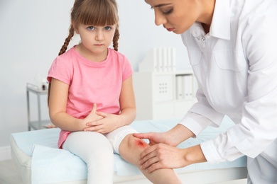 Photo of Female doctor examining little girl's injured leg in clinic. First aid