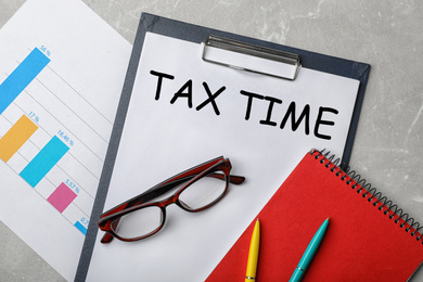Image of Glasses, notebook, chart and paper with words TAX TIME on grey table, flat lay