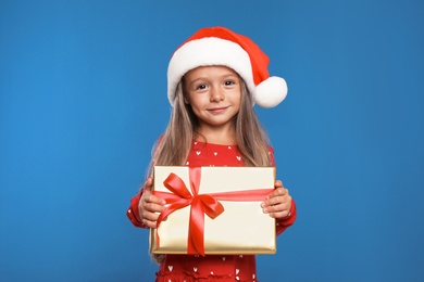Photo of Happy little child in Santa hat with gift box on blue background. Christmas celebration