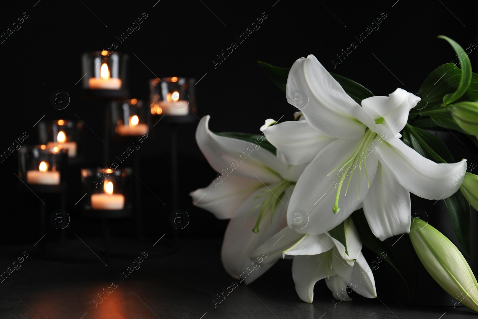 Photo of White lilies and blurred burning candles on table in darkness, closeup with space for text. Funeral symbol