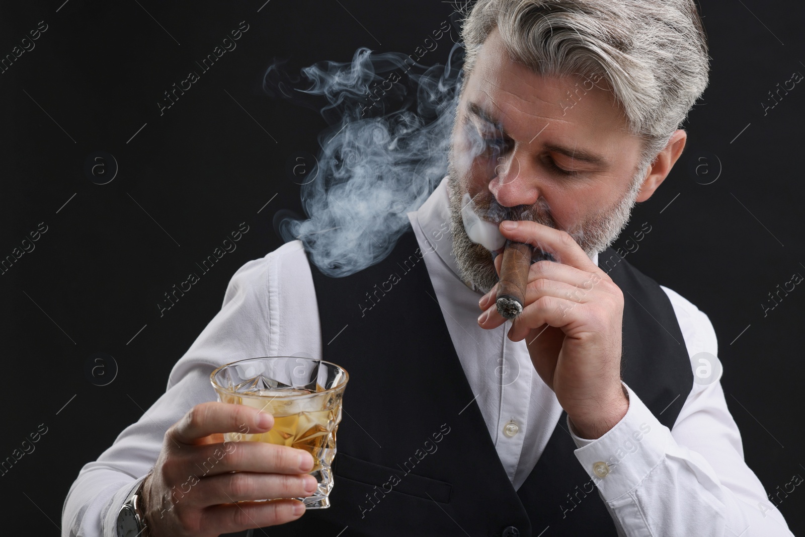 Photo of Bearded man with glass of whiskey smoking cigar against black background