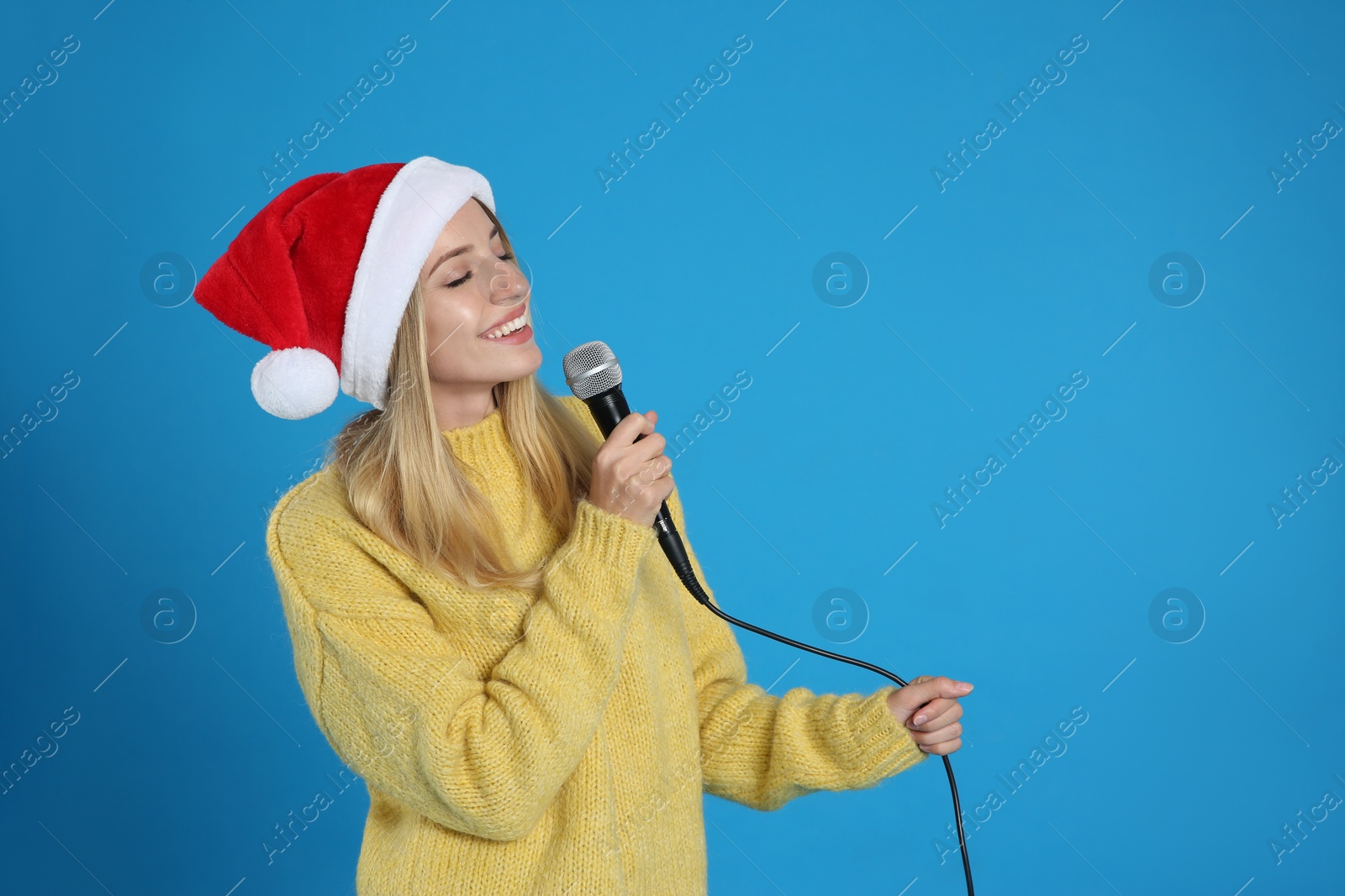 Photo of Happy woman in Santa Claus hat singing with microphone on blue background, space for text. Christmas music