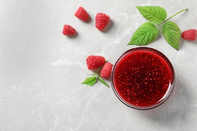 Photo of Bowl of sweet jam with ripe raspberries and green leaves on white table, flat lay. Space for text