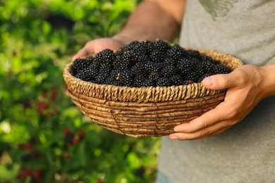 Photo of Woman holding wicker bowl with ripe blackberries outdoors, closeup
