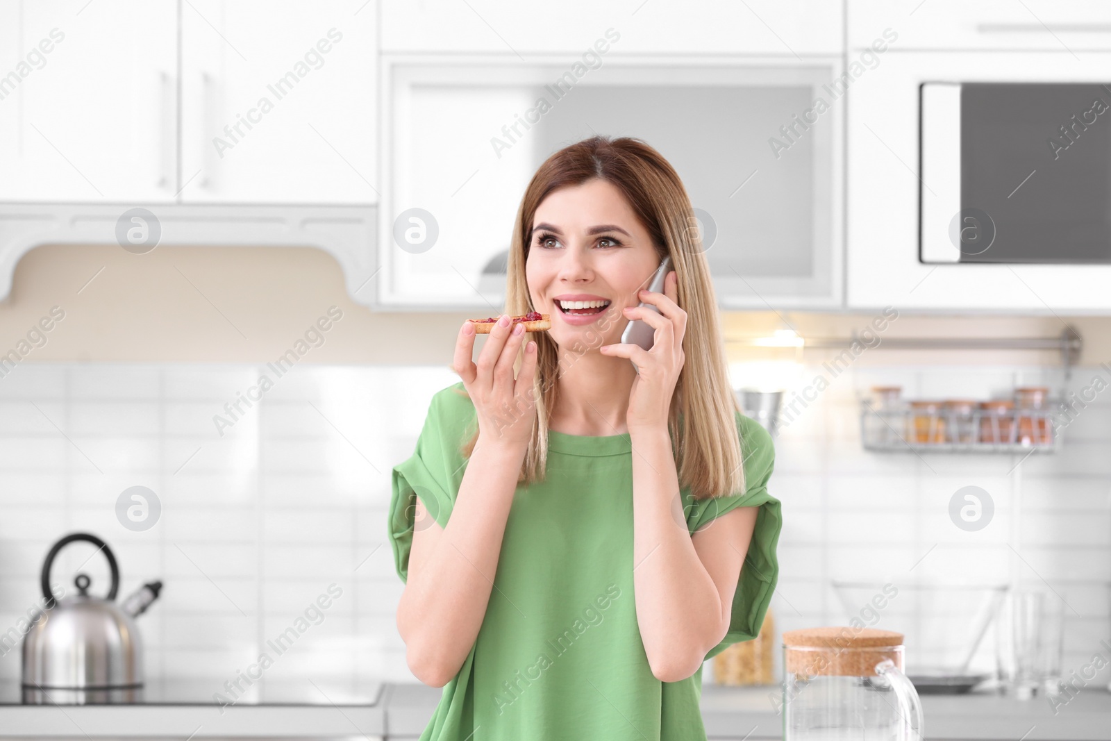 Photo of Beautiful woman eating tasty toasted bread with jam while talking on mobile phone in kitchen