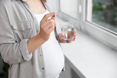 Photo of Pregnant woman holding pill and glass with water near window indoors, closeup