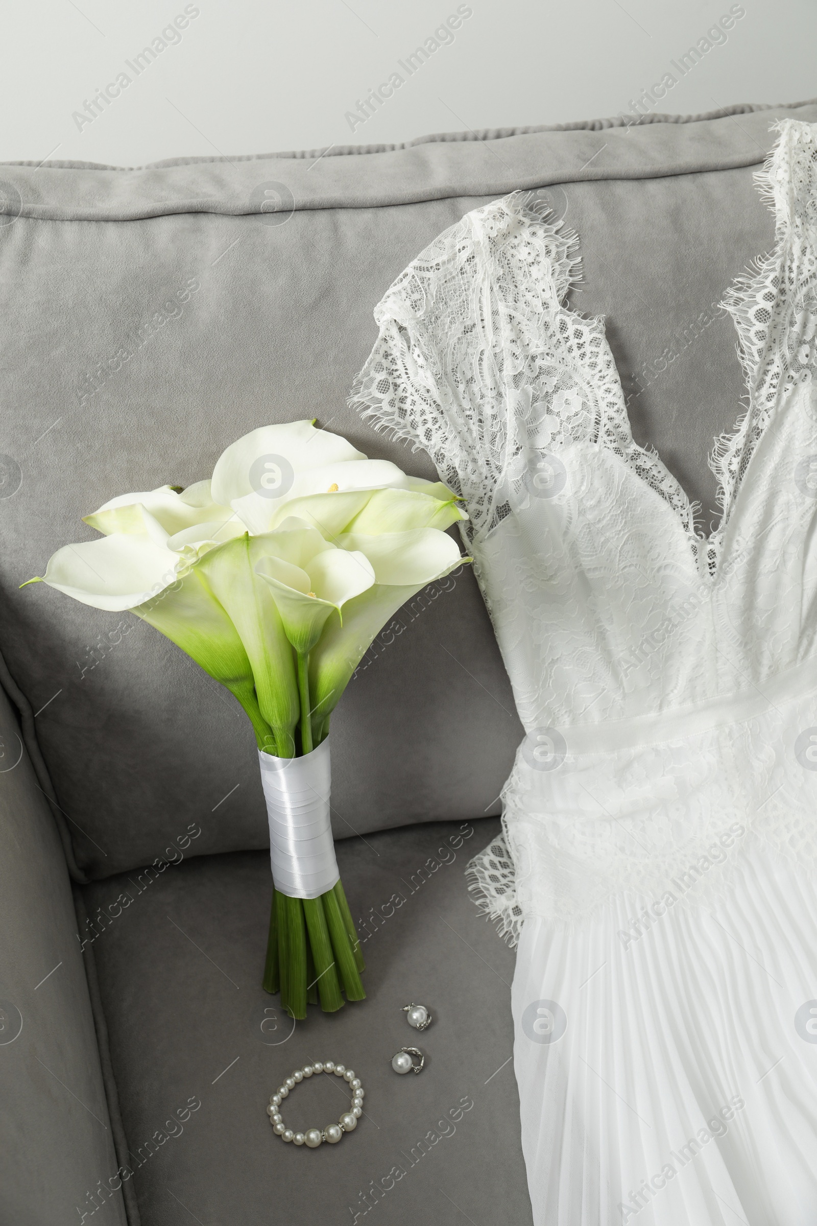 Photo of Beautiful calla lily flowers tied with ribbon, wedding dress and jewelry on sofa