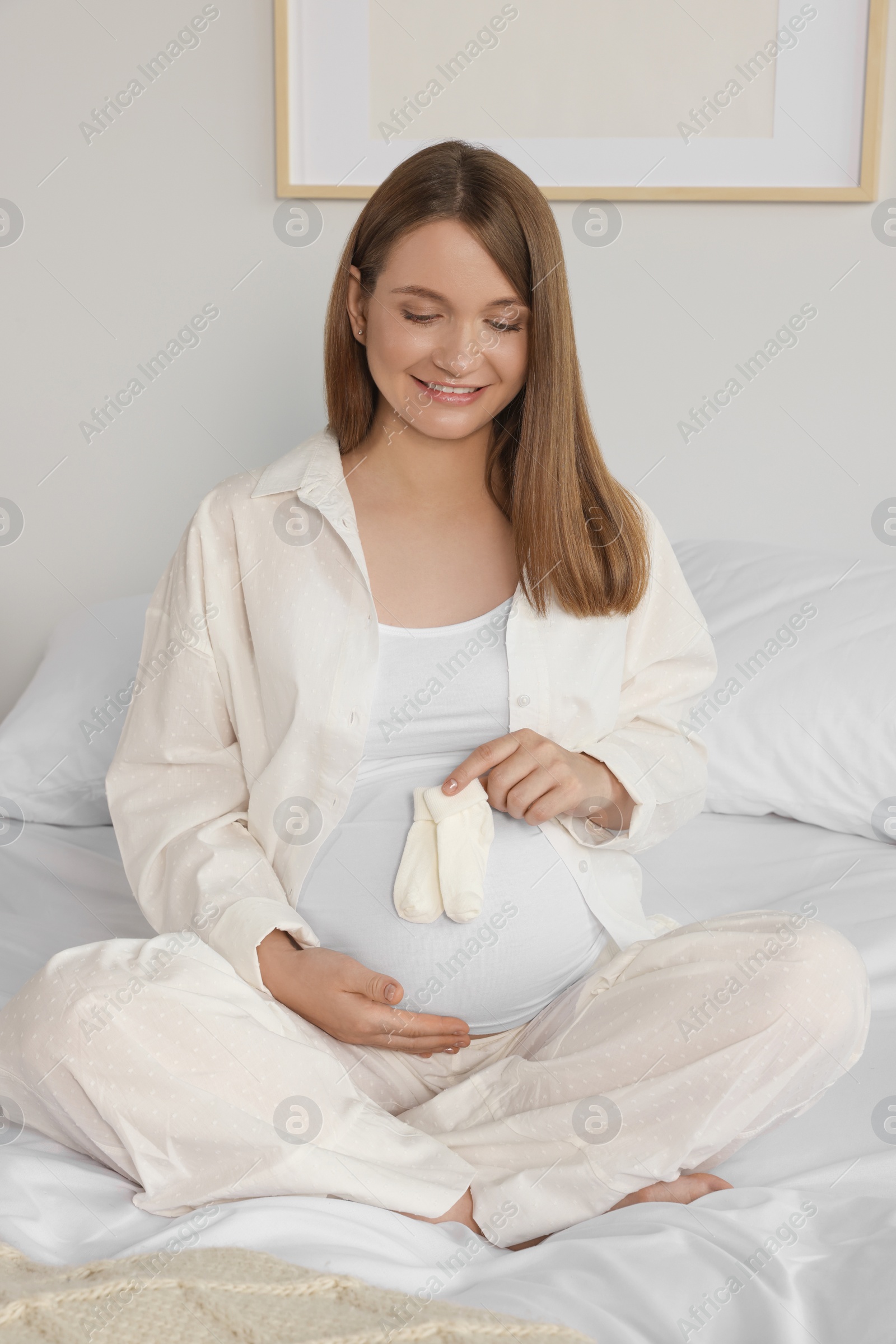 Photo of Happy young pregnant woman with baby socks on bed at home