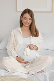 Photo of Happy young pregnant woman with baby socks on bed at home