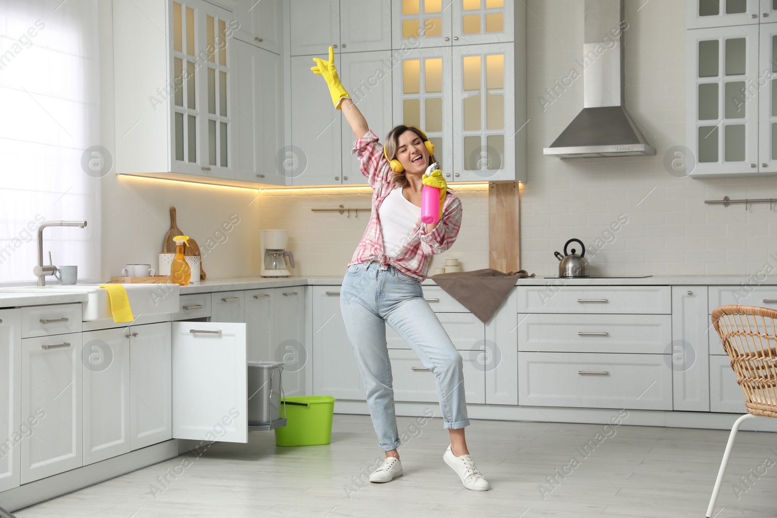 Photo of Beautiful young woman with headphones singing while cleaning kitchen