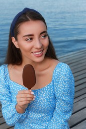 Photo of Beautiful young woman eating ice cream glazed in chocolate on pier