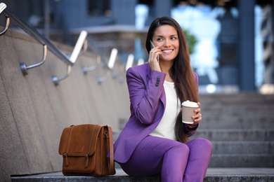 Photo of Beautiful businesswoman with cup of coffee talking on phone outdoors