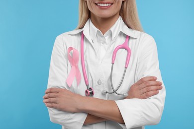 Photo of Doctor with pink ribbon and stethoscope on light blue background, closeup. Breast cancer awareness