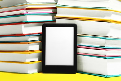 Photo of Modern e-book reader and stacks of hard cover books on yellow background, closeup