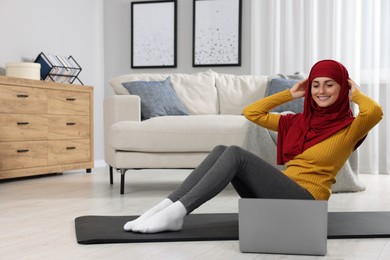 Muslim woman in hijab doing abs exercise near laptop on fitness mat at home. Space for text