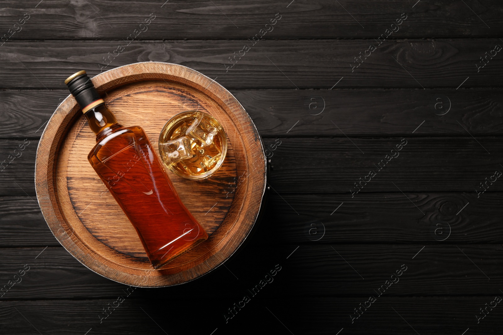 Photo of Whiskey with ice cubes in glass, bottle and barrel on black wooden table, top view. Space for text