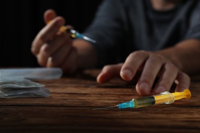 Addicted man at wooden table, focus on syringe. Hard drugs