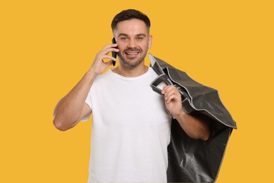 Photo of Man holding garment cover with clothes while talking on phone against yellow background. Dry-cleaning service