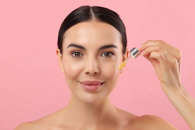 Beautiful young woman applying serum onto her face on pink background