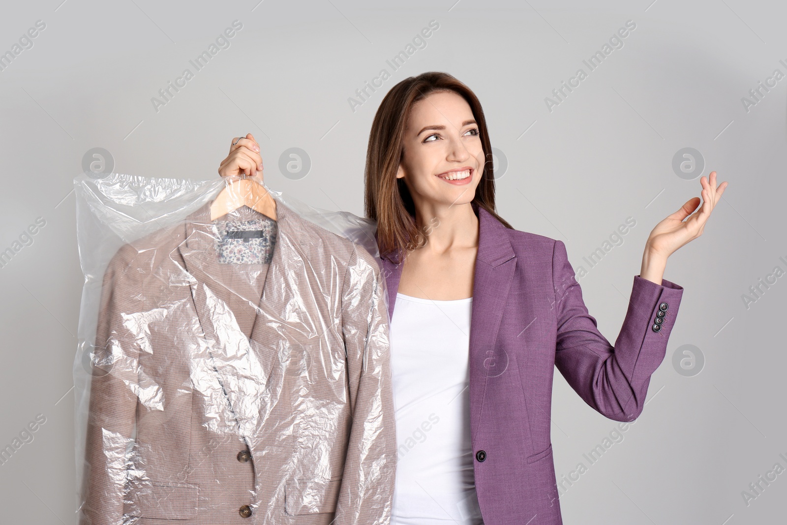 Photo of Young woman holding hanger with jacket in plastic bag on grey background. Dry-cleaning service