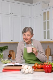 Happy woman with cup of tea and recipe book at table in kitchen