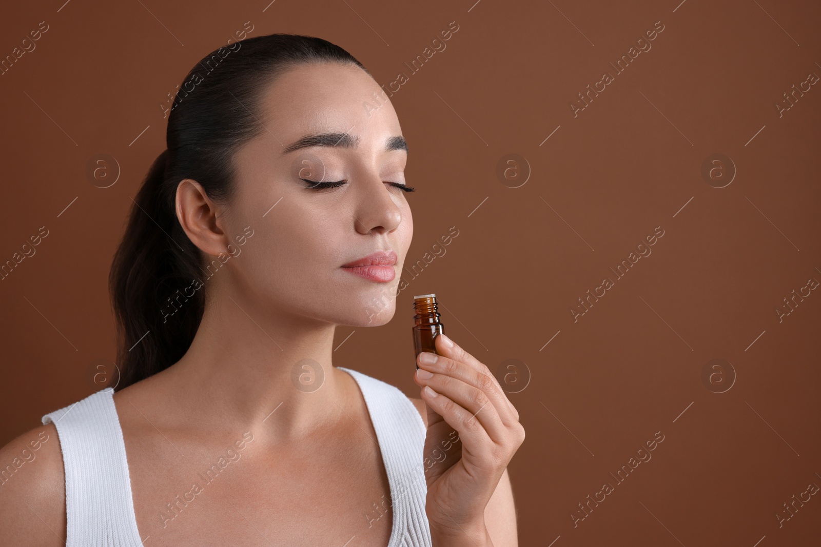 Photo of Beautiful young woman with bottle of essential oil on brown background, space for text