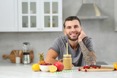 Handsome man with delicious smoothie and ingredients at white table in kitchen. Space for text