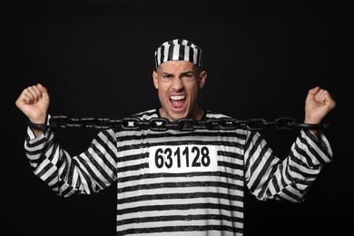 Photo of Emotional prisoner in striped uniform trying to break  chain on black background