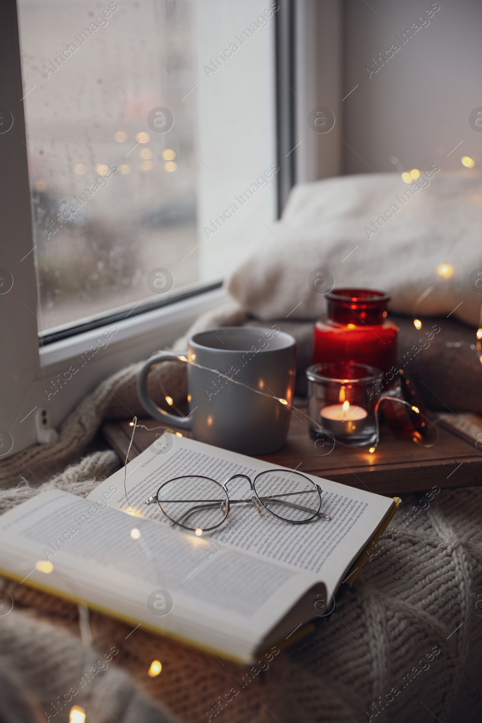 Photo of Cup of hot drink, open book and Christmas lights on knitted blanket near window. Cozy season