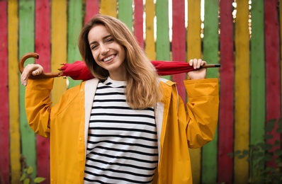 Beautiful young woman in stylish autumn clothes with red umbrella near color fence