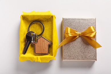 Key with trinket in shape of house and gift box on light grey background, flat lay. Housewarming party