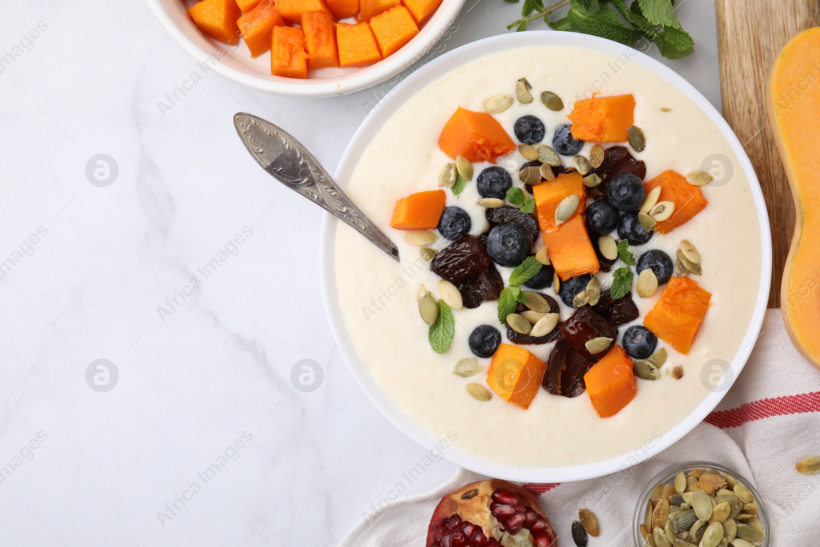 Photo of Bowl of delicious semolina pudding with blueberries, dates, pumpkin, spoon and mint on white marble table, flat lay. Space for text